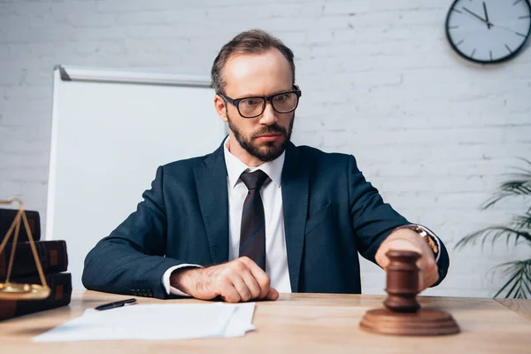 Selective focus of bearded judge in glasses looking at gavel near papers on table — Stock Photo