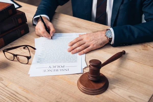 Ausgeschnittene Ansicht des Richters beim Unterschreiben von Papieren mit Versicherungsaufdruck in der Nähe von Hammer und Gläsern auf dem Tisch — Stockfoto