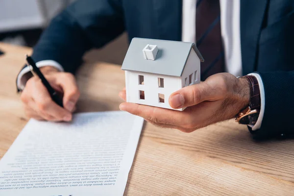 Cropped view of lawyer holding house model while signing documents on table — Stock Photo