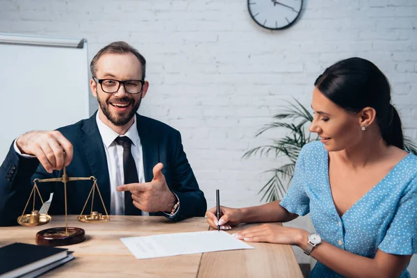 Selektiver Fokus des bärtigen Rechtsanwalts mit Brille, der mit dem Finger auf die Waage zeigt, während die Frau im Büro einen Versicherungsvertrag unterschreibt — Stockfoto