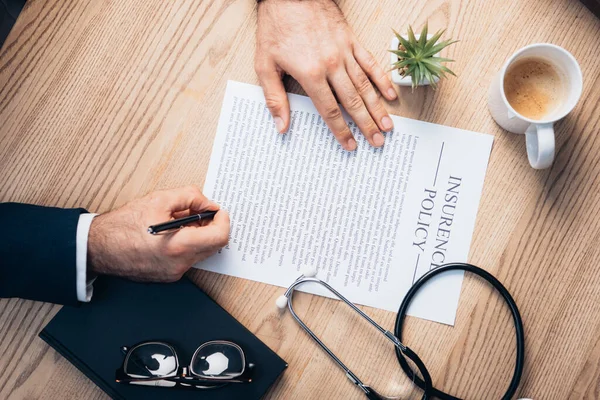 Ausgeschnittene Ansicht eines Rechtsanwalts bei der Unterzeichnung eines Versicherungsvertrages in der Nähe von Werk, Brille, Notizbuch und Stethoskop auf dem Schreibtisch — Stockfoto