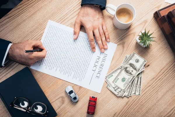 Top view of lawyer signing insurance policy agreement near plant, glasses, notebook, dollars and toy cars — Stock Photo