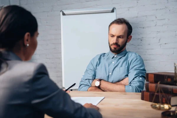 Selektiver Fokus des unzufriedenen Mannes, der mit verschränkten Armen neben Anwalt und Büchern auf dem Tisch sitzt — Stockfoto