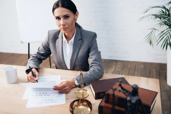 Selective focus of brunette lawyer in formal wear holding pen near insurance policy documents and looking at camera — Stock Photo