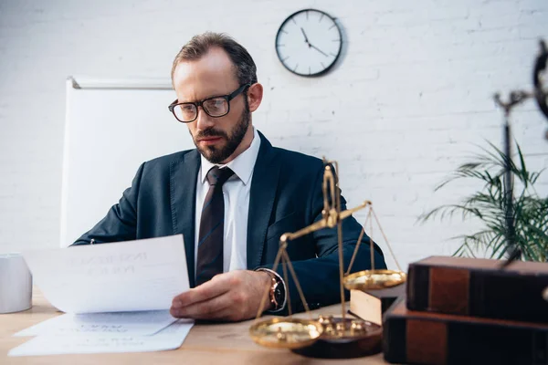 Selective focus of bearded lawyer looking at documents near golden scales and books — Stock Photo