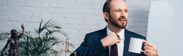 Horizontal crop of lawyer in suit touching tie while holding cup — Stock Photo