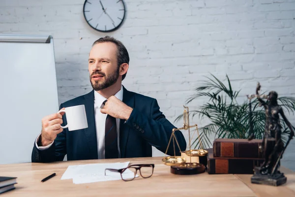 Bearded lawyer in suit touching tie while holding cup in office — Stock Photo