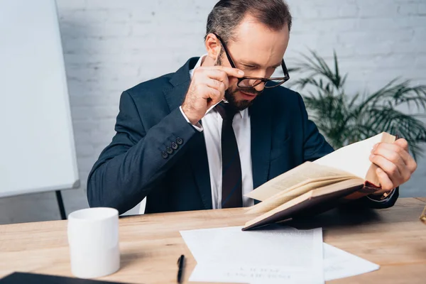 Bärtiger Anwalt im Anzug berührt Brille beim Lesen von Buch im Büro — Stockfoto