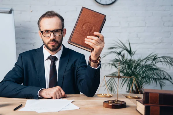 Избирательный фокус бородатого адвоката в очках, держащего книгу в офисе — стоковое фото