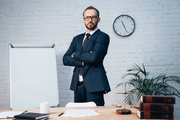 Avocat barbu dans des lunettes debout avec les bras croisés près de la table dans le bureau — Photo de stock