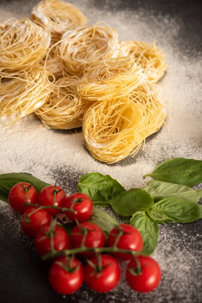 Selective focus of raw Italian Capellini with tomatoes, basil and flour on black background — Stock Photo