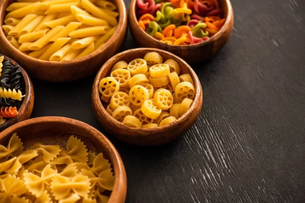 Selective focus of various raw Italian pasta in wooden bowls on black background — Stock Photo