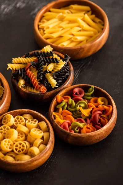 Selective focus of various raw Italian pasta in wooden bowls on black background — Stock Photo