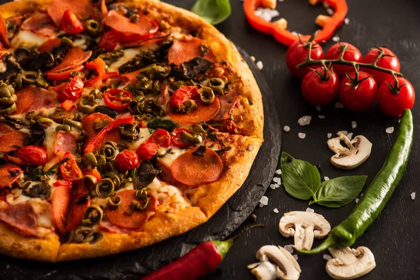 Delicious Italian pizza with salami near vegetables on black background — Stock Photo
