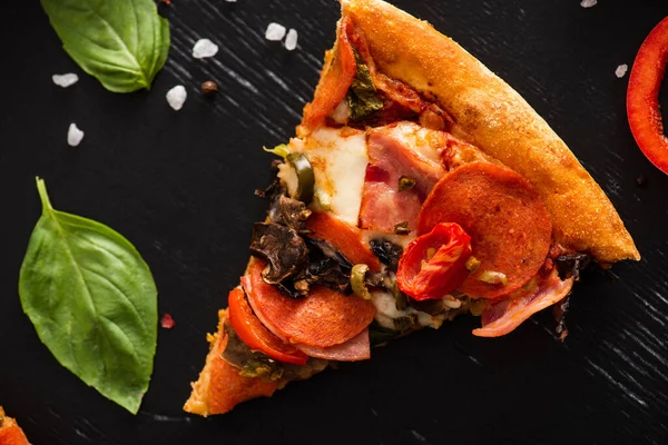 Vista superior de deliciosa fatia de pizza italiana com salame perto de folhas de manjericão e sal no fundo preto — Fotografia de Stock