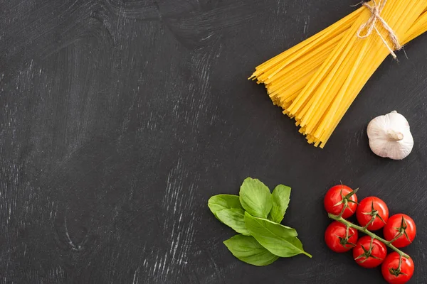 Vista superior de espaguete italiano cru com folhas de manjericão, tomates e alho sobre fundo preto — Fotografia de Stock