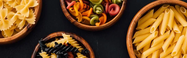 Top view of assorted colorful Italian pasta in wooden bowls on black background, panoramic shot — Stock Photo