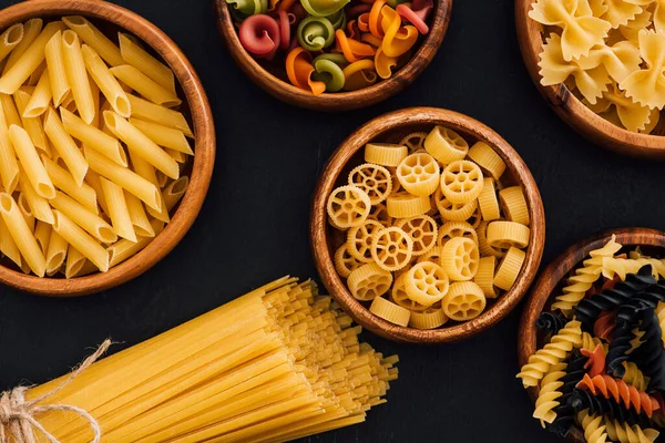 Top view of assorted colorful Italian pasta in wooden bowls on black background — Stock Photo