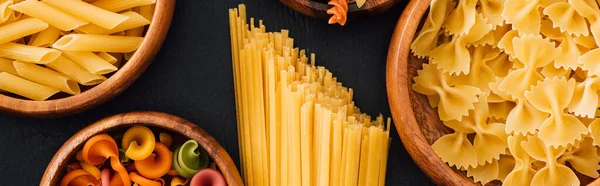 Top view of assorted Italian pasta in wooden bowls on black background, panoramic shot — Stock Photo