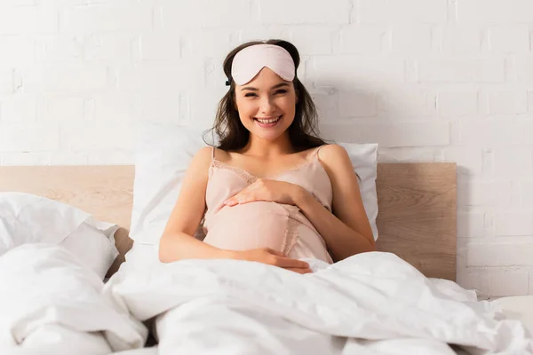 Embarazada joven en camisón de seda tocando vientre en el dormitorio - foto de stock