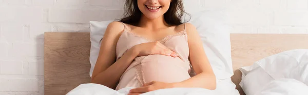 Panoramic crop of pregnant woman in silk nightie touching belly in bedroom — Stock Photo
