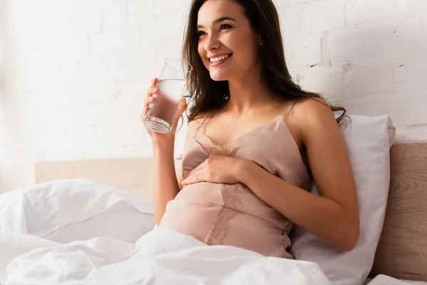 Jovem grávida segurando vidro de água e barriga tocando — Fotografia de Stock