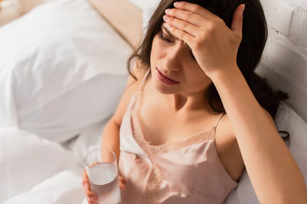 Blick über den Kopf einer müden Frau, die den Kopf berührt und ein Glas Wasser hält — Stockfoto