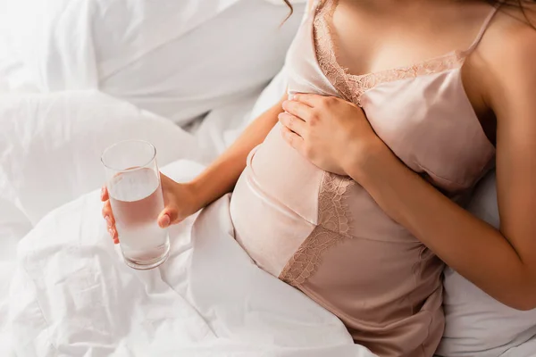 Ausgeschnittene Ansicht einer jungen Schwangeren, die ein Glas Wasser in der Hand hält und den Bauch im Schlafzimmer berührt — Stockfoto
