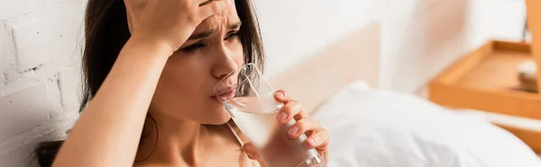 Horizontal image of tired woman touching head and holding glass while drinking water — Stock Photo