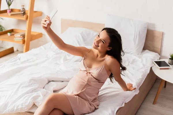 Young pregnant woman sitting on bed and taking selfie — Stock Photo