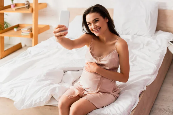 Messa a fuoco selettiva della giovane donna incinta che prende selfie in camera da letto — Foto stock