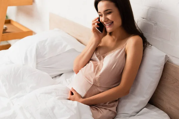Молодая и беременная женщина разговаривает по смартфону в спальне — стоковое фото