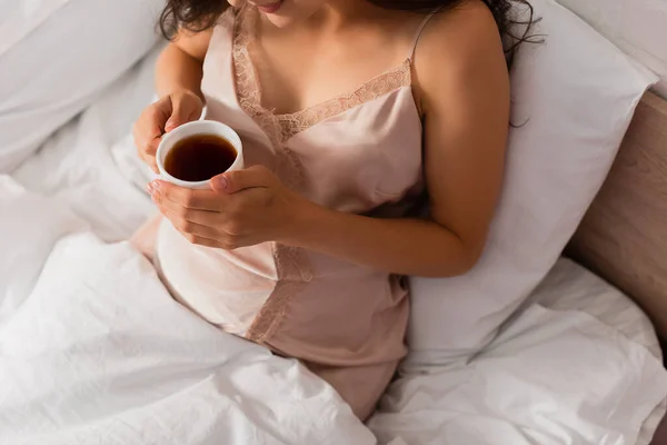 Vista recortada de la mujer en camisón de seda sosteniendo taza de té - foto de stock