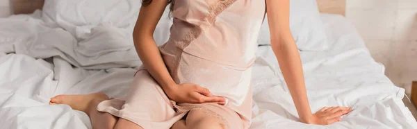 Горизонтальный урожай беременной девушки в шелковой ночнушке, сидящей на кровати — стоковое фото