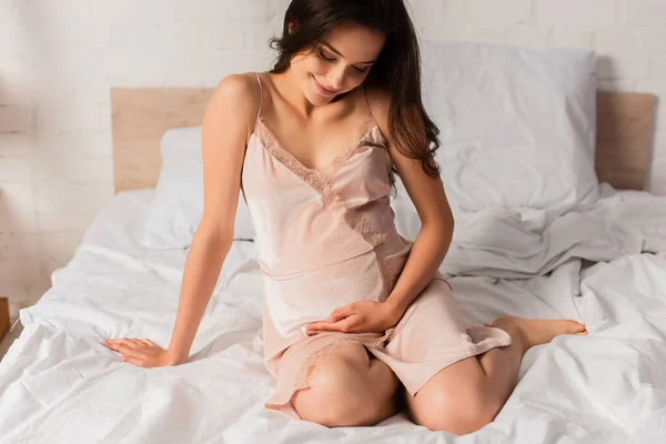 Schwangere junge Frau im seidenen Nachthemd sitzt auf Bett und berührt Bauch — Stockfoto