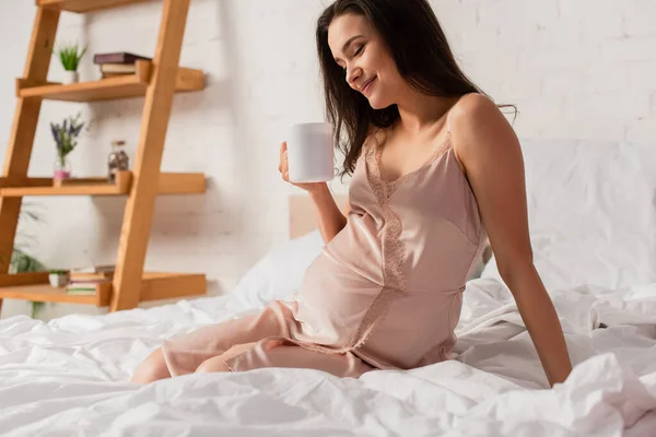 Femme enceinte et brune tenant une tasse de thé au lit — Photo de stock