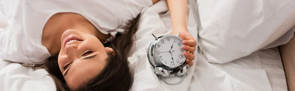 Panoramic shot of young woman lying on bed with retro alarm clock — Stock Photo