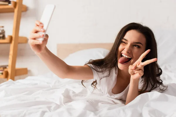 Selektiver Fokus einer jungen Frau, die beim Selfie im Schlafzimmer die Zunge herausstreckt und ein Friedenszeichen zeigt — Stockfoto