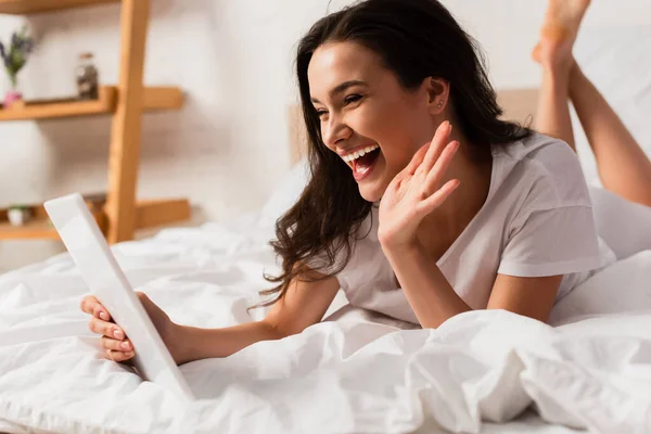 Selective focus of young woman lying on bed, waving hand and holding digital tablet while having video call — Stock Photo