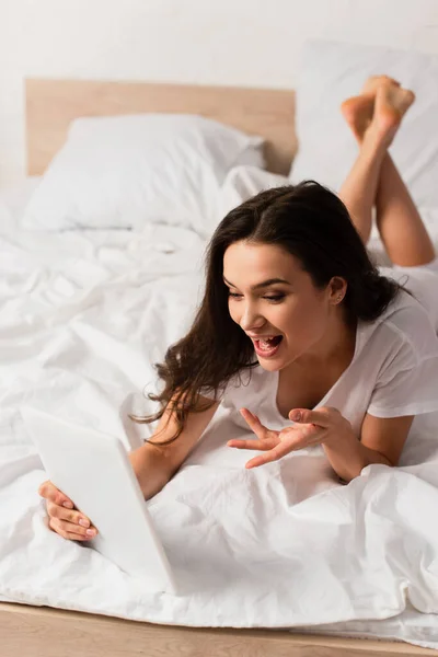 Selektiver Fokus einer jungen Frau, die auf dem Bett liegt und während eines Videogesprächs ein digitales Tablet in der Hand hält — Stockfoto