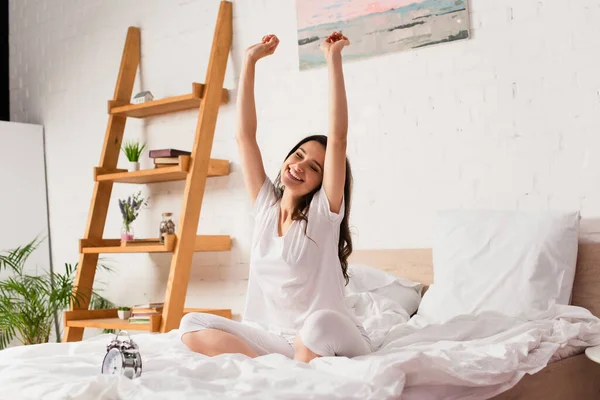Young woman sitting on bed and stretching near retro alarm clock — Stock Photo