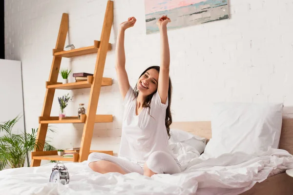 Mujer joven sentada en la cama y estirándose cerca del despertador retro - foto de stock