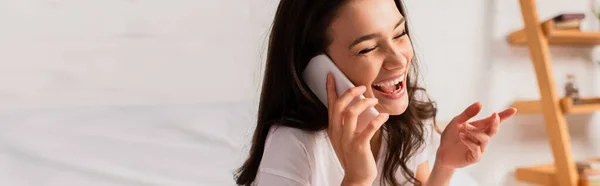 Horizontal crop of brunette woman talking on smartphone while gesturing in bedroom — Stock Photo