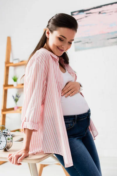 Mulher grávida tocando barriga e de pé perto da mesa com despertador vintage — Fotografia de Stock