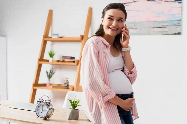 Беременная женщина разговаривает на смартфоне и стоит рядом со столом с винтажным будильником и растением — стоковое фото