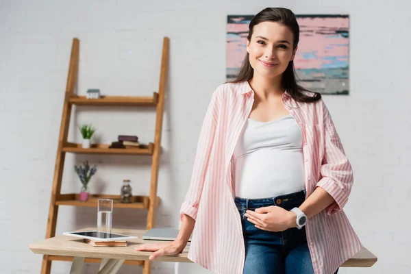 Schwangere berührt Bauch, während sie am Tisch mit Gadgets steht — Stockfoto