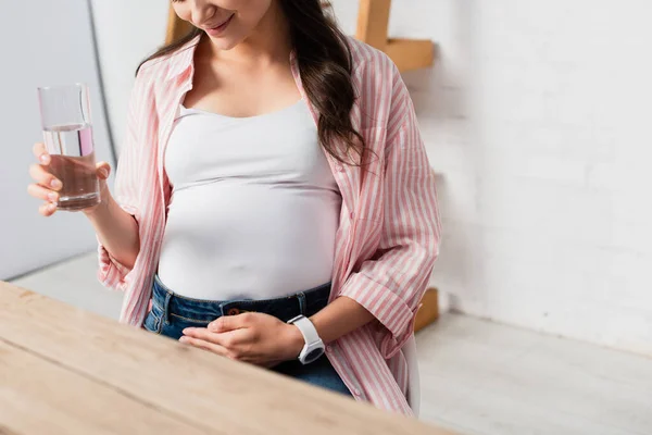Abgeschnittene Ansicht einer schwangeren Frau, die Bauch berührt und ein Glas Wasser in der Hand hält — Stockfoto