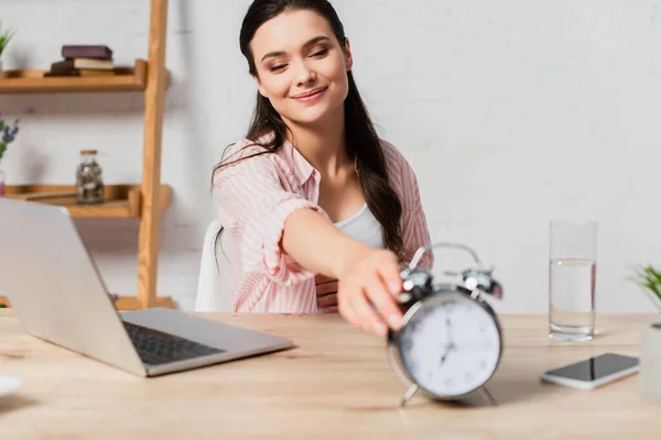 Enfoque selectivo de la mujer morena llegar reloj despertador retro cerca de gadgets en la mesa - foto de stock