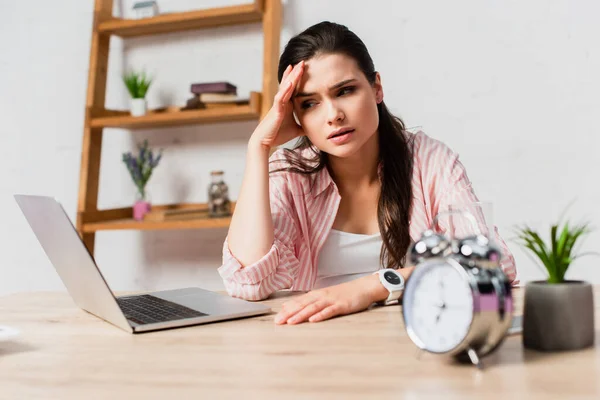 Selective focus of stressed freelancer touching head near alarm clock, plant and laptop — Stock Photo
