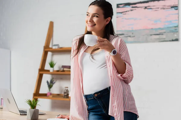 Pregnant woman looking away while holding cup of tea while standing near laptop — Stock Photo